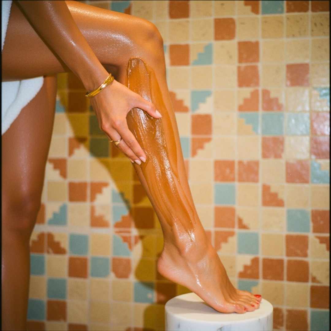 Image éditoriale ultra réaliste d’une femme à la peau marocaine dorée appliquant le Savon Beldi Peau Sensible MARÏYA directement sur sa jambe nue.
La texture noire-brune brillante du savon s’étale sur la peau, évoquant un soin artisanal et apaisant.
En arrière-plan, un mur de carrelage marocain rétro inspire l’ambiance du hammam traditionnel, sublimée par une lumière chaude des années 70.
L’image illustre la richesse, la douceur et la naturalité du rituel corporel marocain signé MARÏYA
