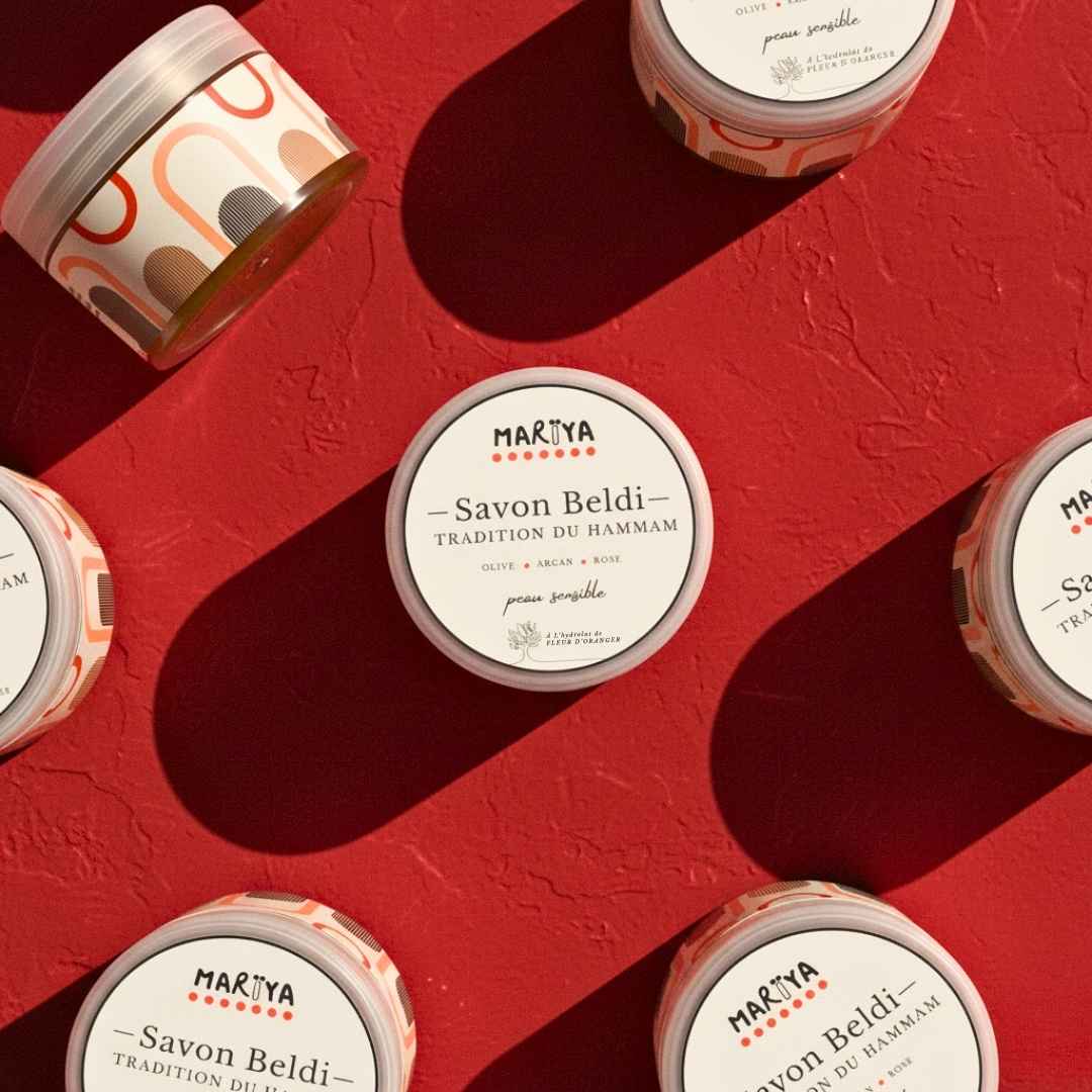 Flatlay éditorial des pots MARÏYA Savon Beldi – Peau Sensible, disposés graphiquement sur un fond rouge grenat vintage, pot central avec étiquette lisible, ombres nettes style rétro.