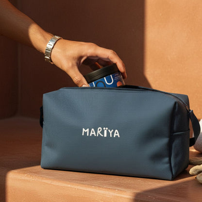 Un plan sur la trousse de bain MARÏYA brodée, posée sur une surface en tadelakt chaud.
L’homme, gourmette au poignet, s’apprête à y glisser son pot ouvert de Savon Beldi Homme.
Autour, serviette blanche et accessoires de hammam créent une ambiance rétro chic.
Un moment suspendu entre artisanat marocain, élégance et transmission.